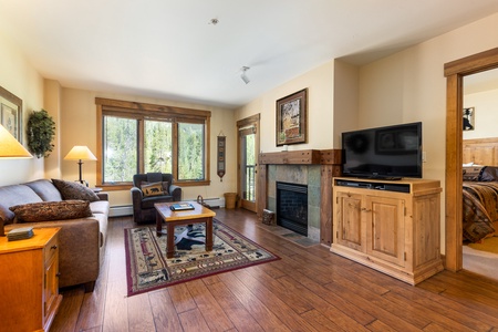 A cozy living room with wood floors, a sofa, a coffee table, an armchair, a TV on a wooden cabinet, and a fireplace. There is a large window with a view and an adjacent doorway to a bedroom.