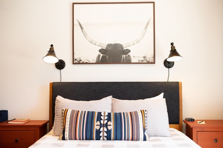 A neatly made bed with a gray headboard, a decorative pillow and bedding, flanked by two bedside tables. A large framed photograph of a longhorn hangs above the bed, with two wall-mounted lamps.