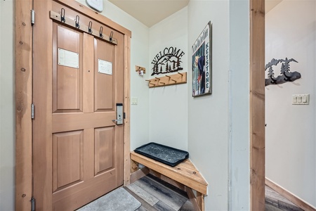 Entrance of a home with a wooden door, coat hooks, a bench, a "Welcome" sign, and a small tray. Light-colored walls and hardwood flooring complete the space.