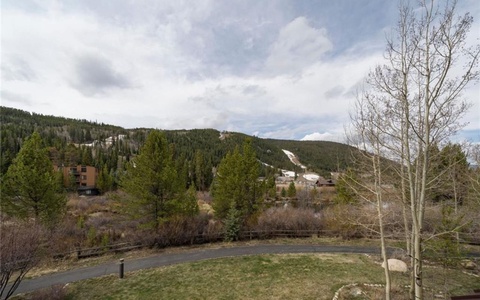 Landscape view of a mountainous area with trees, a winding path, and a few buildings in the distance under a cloudy sky.