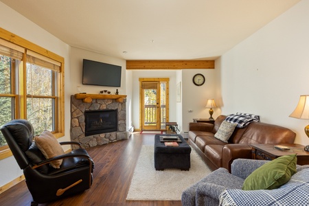 A cozy living room with a leather sofa, an armchair, a stone fireplace, a wall-mounted TV, a wooden coffee table, and large windows showing an outdoor view. A door leads out to a balcony.