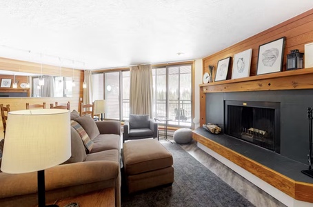 A cozy living room with a beige couch, a gray armchair, and a fireplace. The room has a large window, wooden accents, and framed pictures above the fireplace.