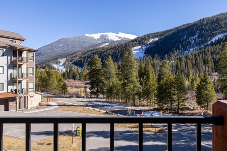 View from a balcony featuring a multi-story building on the left, a paved area below, and a mountainous landscape covered in evergreens and patches of snow in the background.