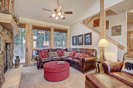 A cozy living room with a brown leather sectional sofa, red ottoman, stone fireplace, and wooden accents. Large windows provide ample natural light, and a staircase is visible in the background.