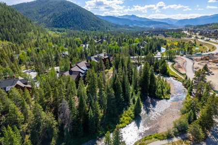 Aerial view of a forested mountain landscape with a winding river, cabins, and distant peaks under a partly cloudy sky.