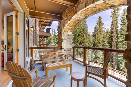 A covered outdoor balcony with wooden seating, a small table, and an archway, overlooks a forested area with tall trees.