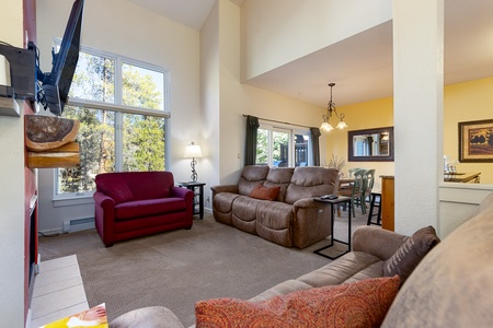 A living room with tall windows, red and brown seating, and a wall-mounted TV. A dining area with a table and chairs is visible in the background, illuminated by a hanging light fixture.