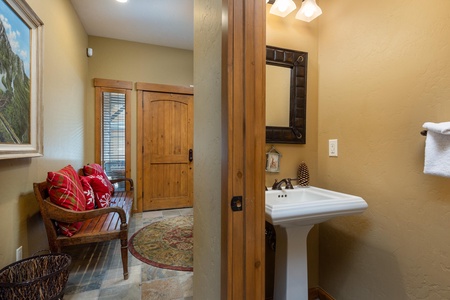 A small bathroom with a pedestal sink and a mirror is adjacent to an entryway featuring a wooden bench with red cushions, a framed painting, and a wooden door.