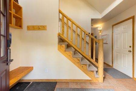 A wooden staircase with a light brown handrail and carpeted steps, adjacent to a white wall in a hallway. Entryway has a small wooden bench and mat near the front door. Wall hooks are mounted nearby.