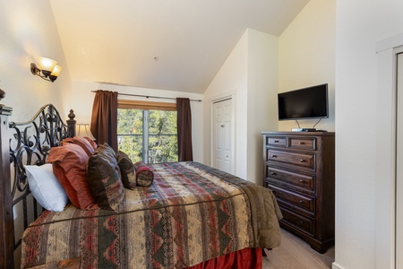 A bedroom with a bed featuring patterned bedding, a wooden dresser with a TV on top, a window with curtains, and beige walls.