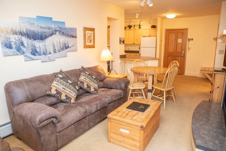 A cozy living room with a brown sofa, wooden coffee table, and round dining table with chairs. The room leads to a kitchen area with wooden cabinets and white appliances. A snowy landscape artwork hangs on the wall.