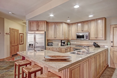 A modern kitchen with wooden cabinets, stainless steel appliances, granite countertops, an island with a sink, and three bar stools. Recessed lighting illuminates the space.