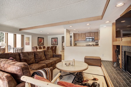 A cozy living room featuring a brown sectional sofa, round coffee table, and fireplace. Adjacent is a kitchen with light wood cabinets and a dining area with a table and chairs. Large windows provide natural light.