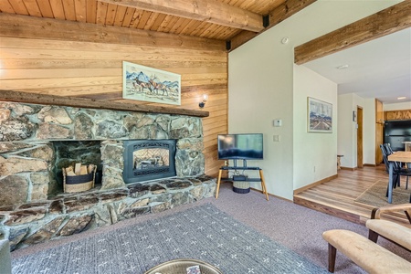 A living room with a stone fireplace, TV, wooden ceiling beams, and a gray patterned rug. The walls have paintings and a hallway leads to a dining area. Logs are stacked by the fireplace.