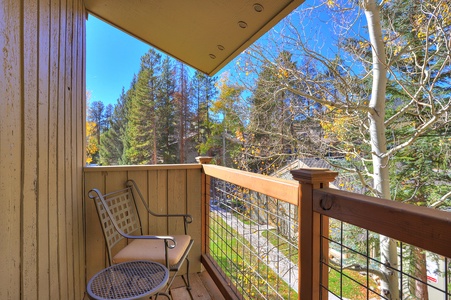 A small balcony features a mesh chair and a round table, overlooking a scenic view of trees and clear blue sky.