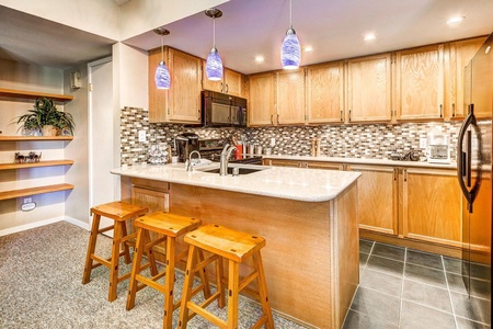 A kitchen with wooden cabinets, a tiled backsplash, an island with a sink, three wooden stools, and three blue pendant lights hanging above.