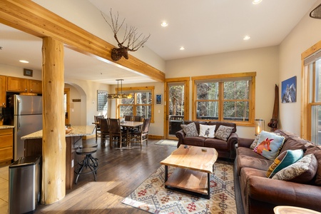 A cozy living room and kitchen area with wooden beams, large windows, a brown leather sofa with colorful pillows, a rustic wooden coffee table, and a dining table with chairs.