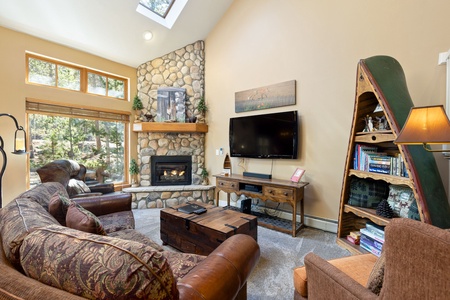 A cozy living room with a stone fireplace, TV, and wooden furniture. The room has a large window, a sofa, a chair, and an open bookshelf shaped like a canoe filled with books and decor.