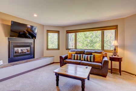 A cozy living room with a wall-mounted TV above a fireplace, a large window with natural light, a brown leather sofa with colorful cushions, a wooden coffee table, and a side table with a lamp.