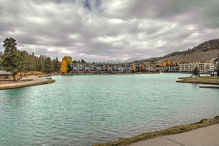 A calm lake with turquoise water is surrounded by apartment buildings, some trees with autumn foliage, and mountainous terrain under a cloudy sky.