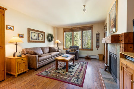 A cozy living room with a sofa, armchair, wooden furniture, and a fireplace. Large windows provide a view of trees outside. The room is decorated with framed pictures, a wreath, and a patterned rug.