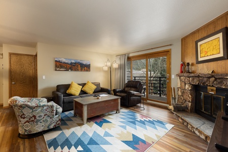 A cozy living room featuring a sectional sofa with yellow pillows, an armchair, a patterned chair, a wooden coffee table, a fireplace with a stone surround, and a colorful geometric rug.