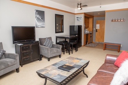 A furnished living area featuring a leather sofa, armchairs, a coffee table, a TV on a stand, a dining set, a kitchenette, and a hallway leading to other rooms. Neutral decor with wooden accents.