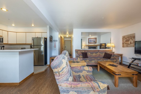 A living room with patterned couches, a wooden coffee table, a TV, and a kitchen area with white cabinets and stainless steel appliances in the background.