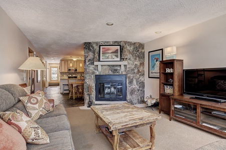 A cozy living room with a stone fireplace, wooden coffee table, sofa with patterned pillows, TV on a wooden stand, and an open view of the kitchen in the background.