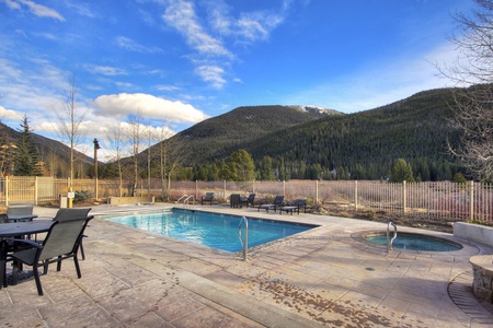 Outdoor swimming pool and hot tub on a concrete deck, surrounded by lounge chairs and tables, set against a backdrop of forested mountains under a clear blue sky.