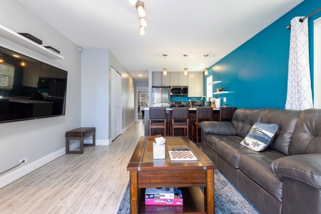 Modern living room with gray couch, wooden coffee table, board games, wall-mounted flat-screen TV, and view into dining area and kitchen with blue accent wall and hanging lights.