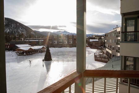 A sunlit view of an outdoor ice skating rink with a Christmas tree at the center. Two people are skating on the rink, and buildings and snow-covered mountains are visible in the background.