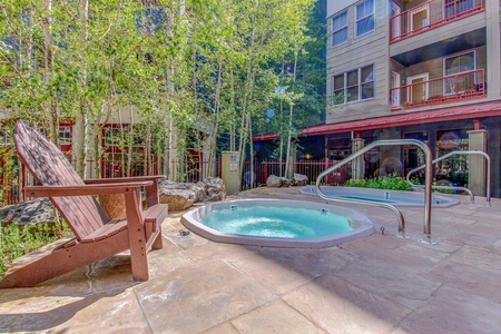 Outdoor hot tub area with surrounding trees and apartment buildings in the background. Patio chair is nearby on stone-paved flooring.