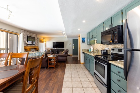 A modern open-concept kitchen and living area with green cabinets, stainless steel appliances, a wooden dining table, hardwood floors, and a wall-mounted TV.