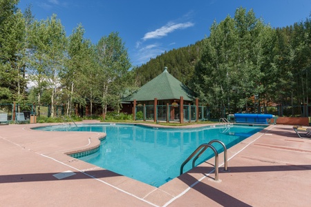 Outdoor swimming pool surrounded by trees with a green-roofed pavilion and mountains in the background. The pool has a ladder and is adjacent to a beige pool deck.