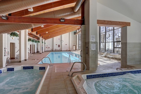 An indoor pool area with natural light, featuring a large swimming pool, two hot tubs, and lounge chairs alongside floor-to-ceiling windows. Wood beams accent the ceiling.