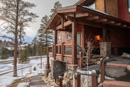 A wooden cabin entrance features a stone foundation and steps, set amidst a snowy landscape with pine trees and a nearby road.