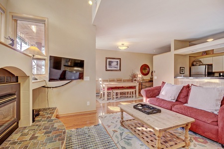 Living room with a red couch, wooden coffee table, wall-mounted TV, and fireplace, opens to a dining area with a long table and a kitchen with stainless steel appliances.