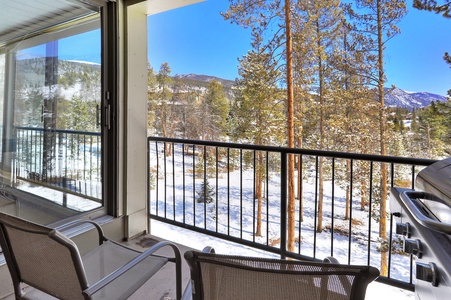 A balcony with two chairs, a barbecue grill, and a scenic view of snow-covered trees and distant mountains on a clear day.