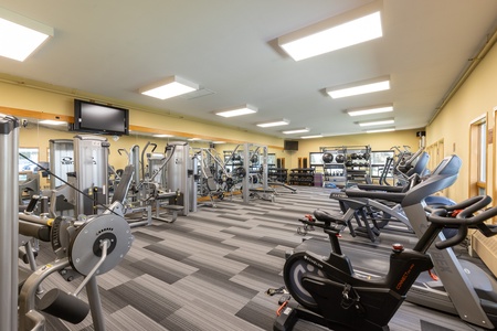 A modern gym with various exercise machines including treadmills, stationary bikes, weight machines, and free weights, well-lit with overhead lighting.