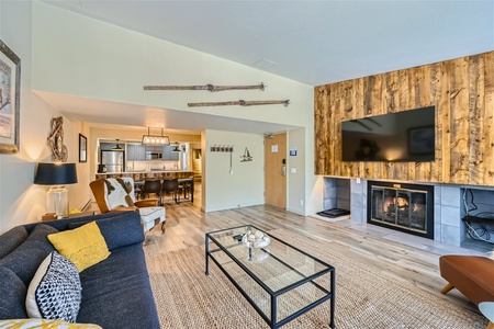 A cozy living room features a fireplace, wall-mounted TV, and rustic decor with wood paneled accents and ski-themed decorations. A kitchen with modern appliances is visible in the background.