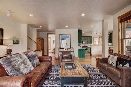 A living room with brown leather sofas, a wooden coffee table, and a patterned rug. The room opens to a kitchen with green cabinetry and a wooden dining table. Stairs lead to an upper floor.