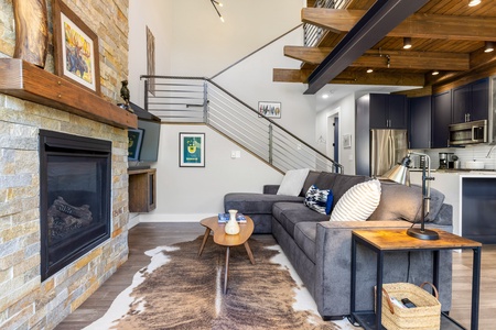 Modern living room with a gray sofa, coffee table, and fireplace. Open kitchen with stainless steel appliances. Exposed beams and a staircase lead to the upper level. Rug on the wooden floor.