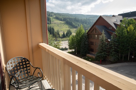 A view of a mountainous landscape from a wooden balcony with a black metal chair. The scene includes a large cabin-style building surrounded by trees.