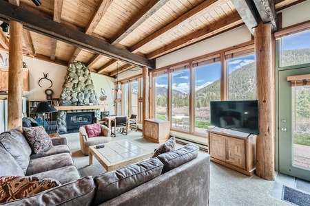 A cozy cabin living room with wooden beams, large windows showcasing a scenic mountain view, a stone fireplace, a sectional sofa, a wooden coffee table, and a TV on a stand.