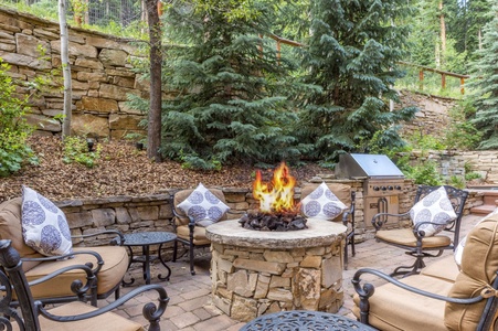 An outdoor patio with a stone fire pit encircled by cushioned chairs. A grill station is on the right, and the area is surrounded by stone walls and tall trees.