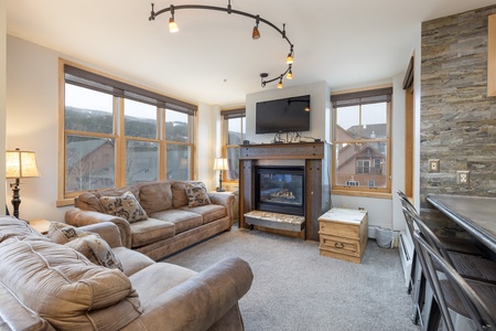 A cozy living room with two beige sofas, a stone fireplace with a TV mounted above it, large windows offering a mountain view, and a bar counter with stools.