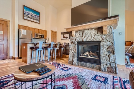 A cozy living space with a stone fireplace, flat-screen TV, and a wooden coffee table. The room features a modern kitchen with bar seating, colorful rugs, and mountain-themed wall art.