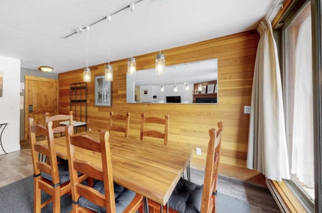 A dining room with a wooden table and chairs, a pendant light fixture, wood-paneled walls, a large mirror, and a sliding glass door with curtains.