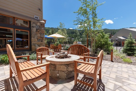 Outdoor seating area with wooden chairs around a stone fire pit on a paved patio, surrounded by greenery and trees, with a building and mountains in the background on a sunny day.